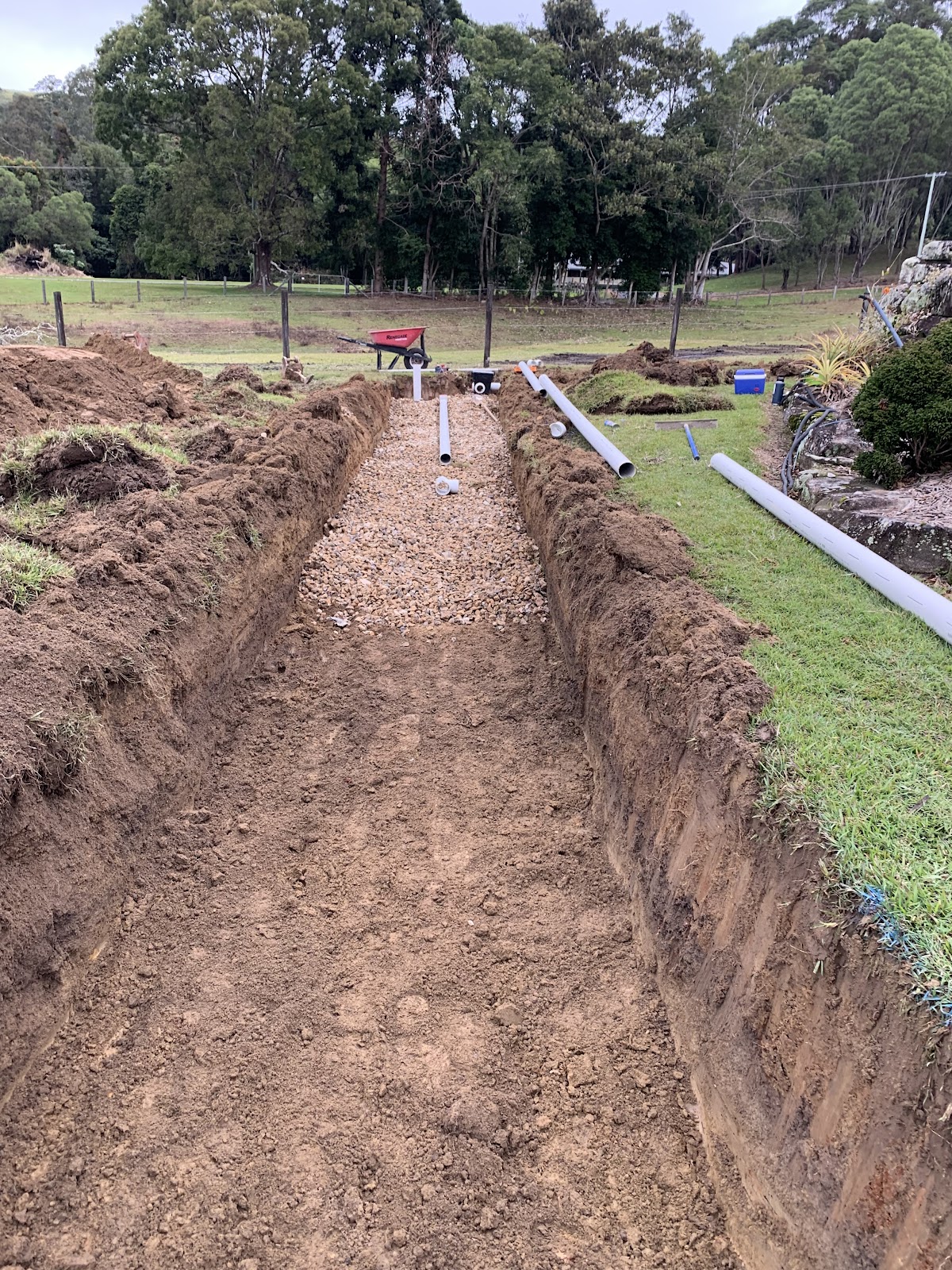 A long gravel-lined trench with pipes on a rural property with trees behind